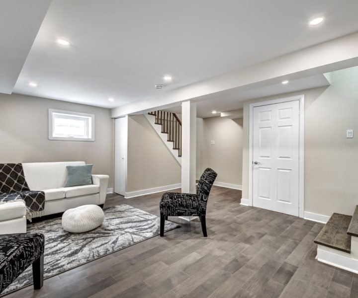 Bright, modern basement living space with dark gray plank flooring, white sectional sofa, patterned rug and chairs, recessed lighting, and a white door—highlighting functional design and seamless integration with upper levels.