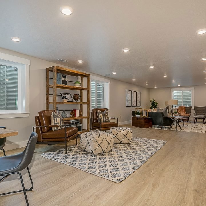 Spacious, fully finished basement with light-toned engineered wood flooring, recessed lighting, multiple egress windows, a dining table with industrial chairs, leather armchairs, patterned rugs, and open shelving—demonstrating seamless, contemporary design integration.