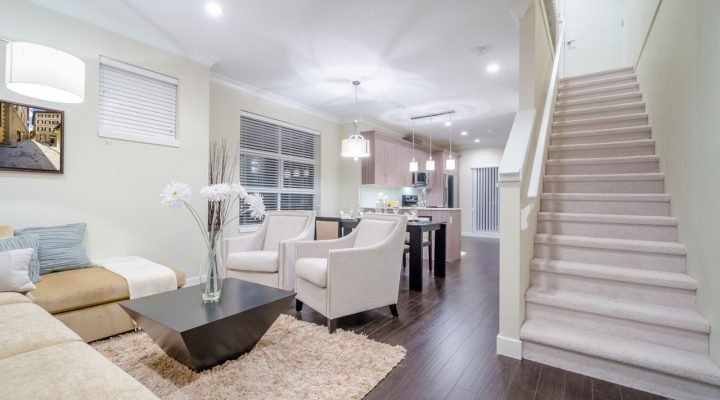 Seamless transition from living room to kitchen in a remodeled lower level: beige carpeted stairs with white railing, cream walls, dark hardwood floors, modern furniture, large windows with blinds, and pendant lighting—demonstrating integrated, high-end renovation.