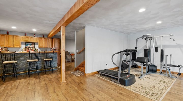 Finished basement featuring a rustic stone-and-wood bar with stools, exposed wood beam ceiling, light hardwood flooring, and a dedicated fitness zone with treadmill and weight machine on an ornate rug—showcasing versatile, high-end remodeling.