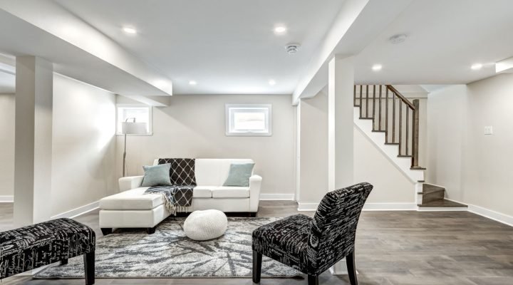 Spacious, elegantly finished basement with gray wood-look flooring, white L-shaped sectional, black-and-white patterned armchair and ottoman, layered rug, two small windows, and a rustic-modern staircase—highlighting cohesive design and comfort.