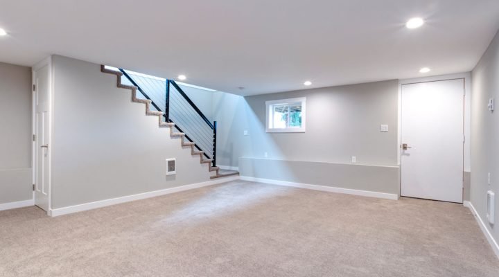 Clean, unfurnished basement with beige carpet, light gray walls, recessed ceiling lights, a small egress window, and a contemporary staircase with black metal railing and—ready for customization by Teton Repair & Remodel.