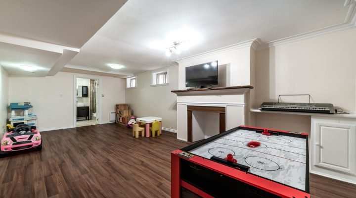 Bright, family-oriented basement play area with dark wood-look flooring, red air hockey table, mounted TV above a white fireplace mantel, pink ride-on car, colorful kids’ furniture, and built-in shelving—designed for fun and functionality.