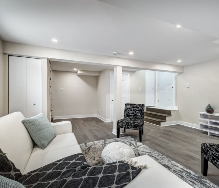 Stylishly finished basement featuring gray wood-look flooring, white sofa with patterned throw, black-and-white accent chair, recessed ceiling lights, and a clean, open layout with storage closet and stair access.