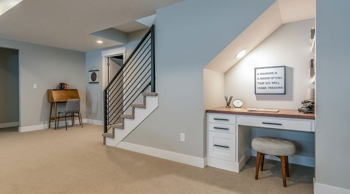 Efficiently designed study nook in a finished basement: white built-in desk with wood top, gray upholstered stool, framed inspirational quote (“A diamond is a chunk of coal that did well under pressure”), and modern black metal staircase nearby.