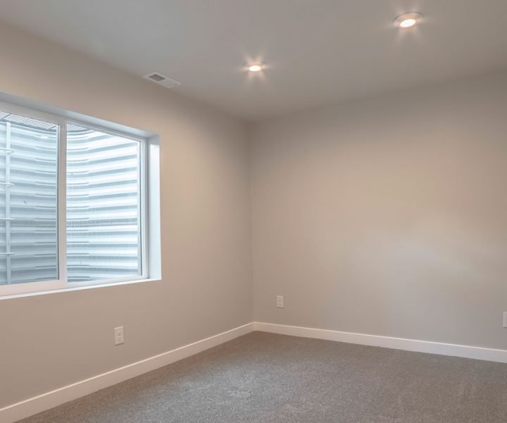 Clean, unfurnished basement room with beige carpet, light gray walls, white baseboards, recessed ceiling lights, and a single egress window with horizontal blinds—ready for customization or final staging.