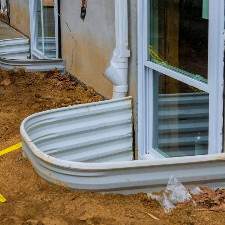 Two newly installed basement egress windows with galvanized steel well covers, set into exposed foundation walls; surrounding dirt, yellow caution tape, and downspout indicate active construction phase—emphasizing code-compliant safety upgrades.