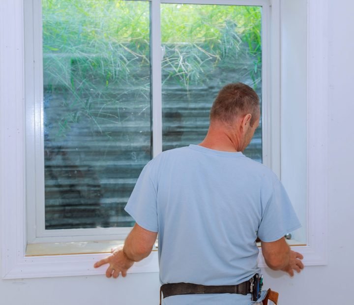 A Teton Repair & Remodel technician in a light blue shirt and tool belt carefully fits a white vinyl egress window into an interior wall, with exterior greenery visible through the glass—highlighting professional installation process.