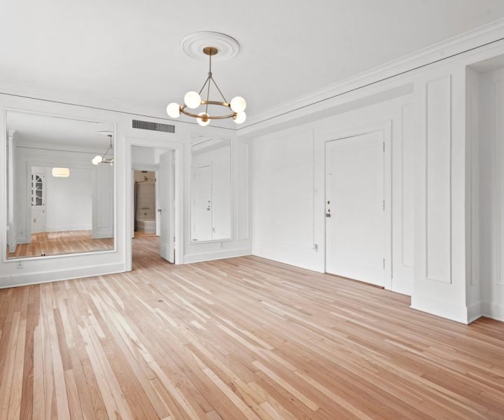 Spacious, empty room with light-toned hardwood flooring, white walls and trim, a modern brass-and-glass chandelier, large mirrors, and a built-in cabinet—showcasing high-quality flooring work by Teton Repair & Remodel.