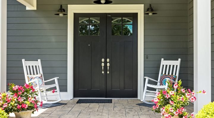 Welcoming front entrance featuring charcoal-gray siding, black double doors with glass panels, white rocking chairs with colorful pillows, potted pink flowers, and a stamped concrete floor—showcasing curb appeal and exterior refinement.