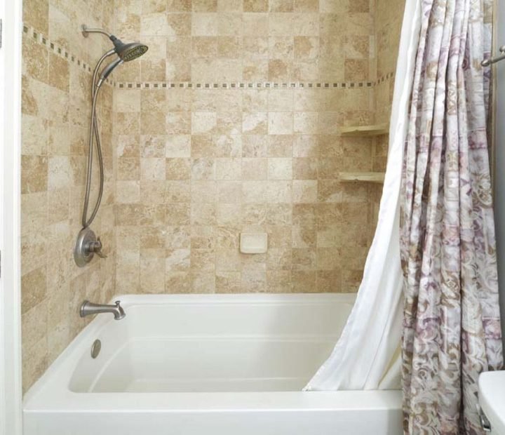 A bathroom featuring a white bathtub with an integrated shower, tiled in beige stone-look squares with a decorative mosaic border. A patterned shower curtain is partially drawn, and built-in shelves are visible in the shower wall.