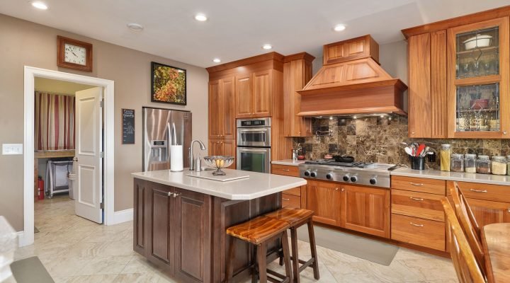 A spacious kitchen with medium-toned wood cabinets, a large dark wood island with bar stools, stainless steel appliances, and a tiled backsplash. A doorway leads to another room.