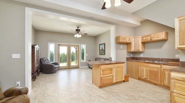 An open-concept living room and kitchen area with light beige tiled floors, light wood cabinetry, and a central island. Large glass doors in the background lead to an outdoor space.