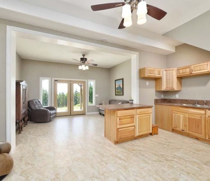An open-concept living room and kitchen area with light beige tiled floors, light wood cabinetry, and a central island. Large glass doors in the background lead to an outdoor space.