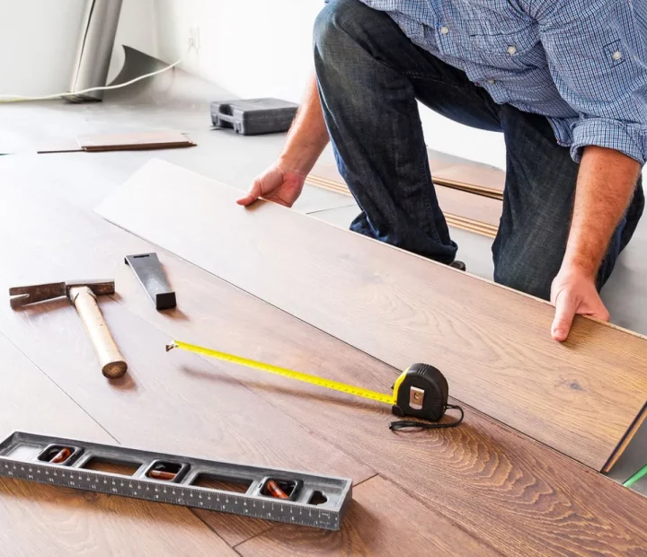 A person is installing laminate or hardwood flooring, kneeling on the floor with a plank in hand. Tools including a hammer, tape measure, and level are laid out on the partially installed floor.