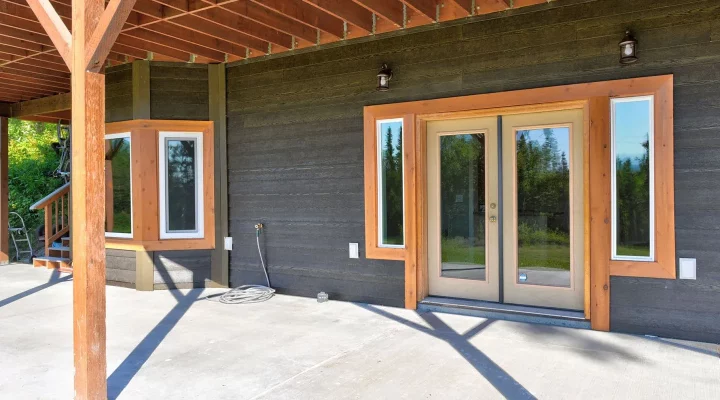 An unfinished covered patio with exposed wooden beams and posts, concrete flooring, and a house exterior featuring dark gray horizontal siding and large windows with wood trim.