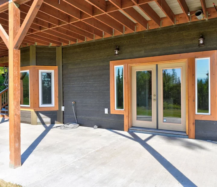 An unfinished covered patio with exposed wooden beams and posts, concrete flooring, and a house exterior featuring dark gray horizontal siding and large windows with wood trim.