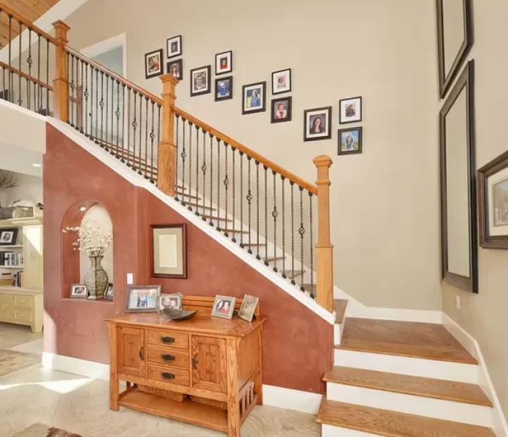 A spacious foyer with a wooden staircase featuring ornate iron balusters, a reddish-brown accent wall, and a gallery wall of framed photos above a wooden console table.