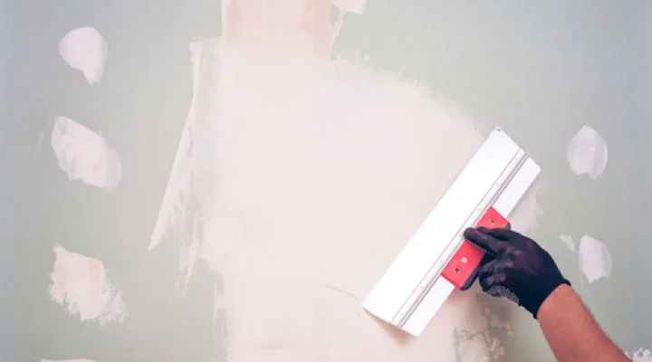A gloved hand uses a wide putty knife to smooth white plaster onto a light green wall, with several other patched areas visible.