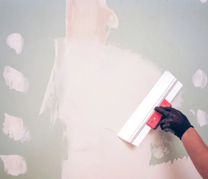 A gloved hand uses a wide putty knife to smooth white plaster onto a light green wall, with several other patched areas visible.