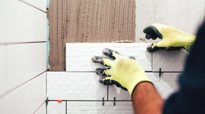 Close-up of hands in yellow gloves setting white textured ceramic tiles on a wall; thinset mortar applied with notched trowel, tile spacers and leveling clips ensure uniform grout lines—demonstrating expert tile installation technique.